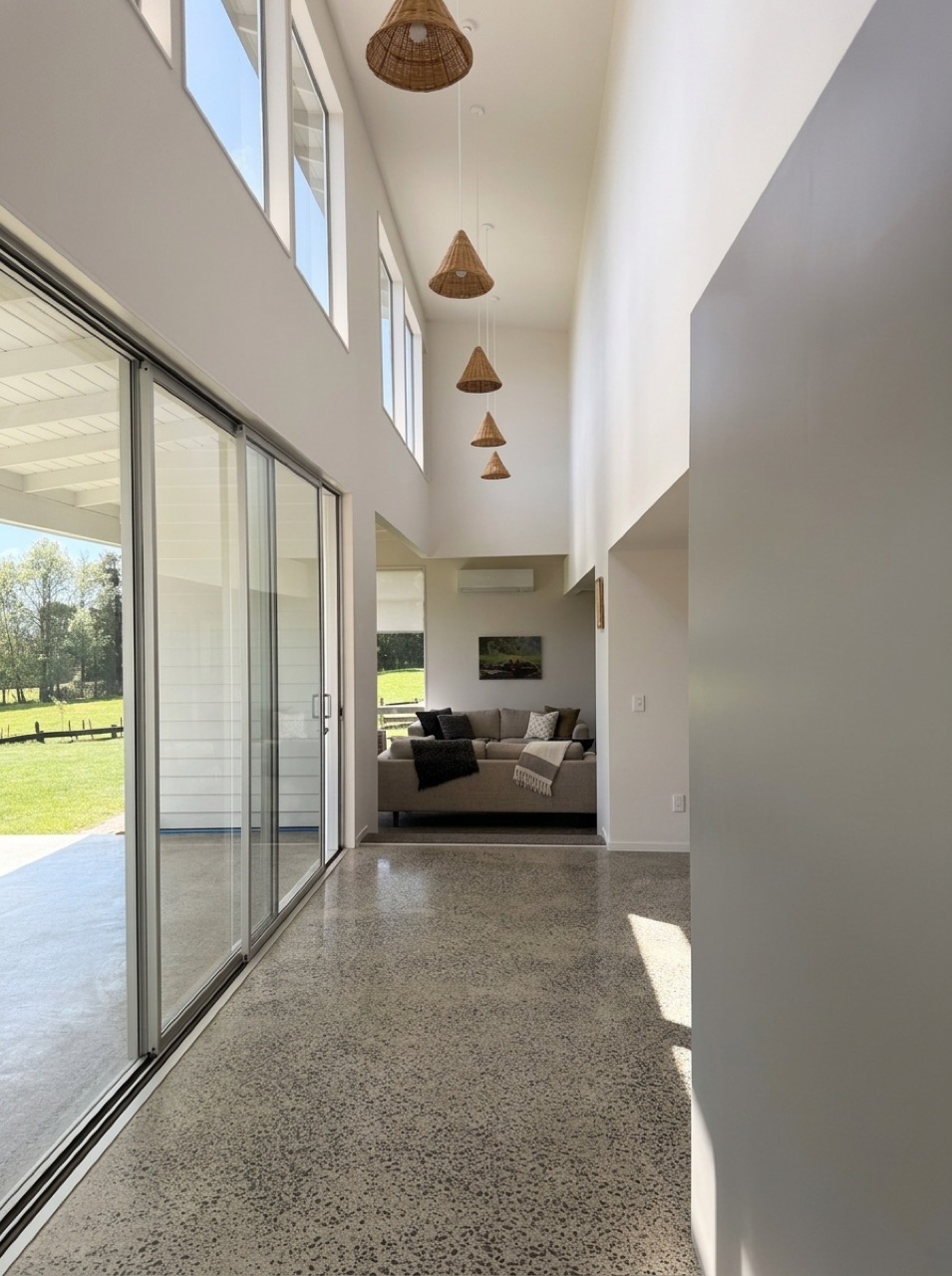 Double-height gallery hallway in natural daylight with polished terrazzo floor, woven rattan pendants, and garden view