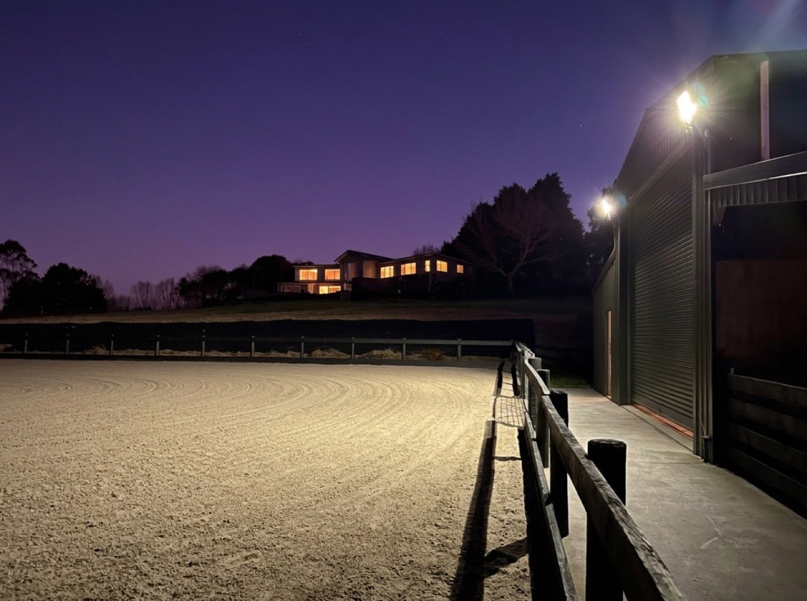 393 m² architectural homestead glowing warmly at night above the freshly groomed floodlit sand arena and barn