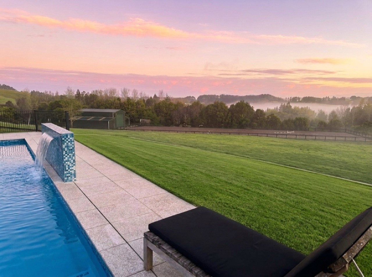 Heated swimming pool edge, flowing blue mosaic waterfall wall, stone terrace, and sun lounger overlooking the misty valley at pink sunrise