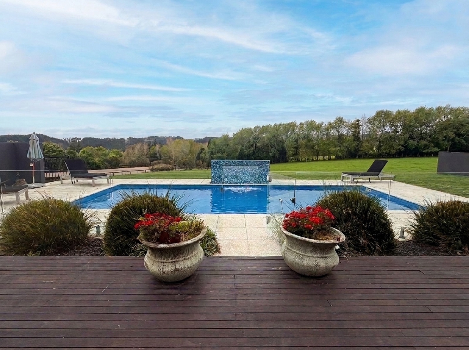 Daytime view from the timber deck across the 21 kW heated swimming pool and cascading blue mosaic waterfall feature to the green hills